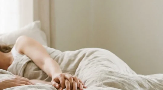 Couple adulte allongé sous des draps clairs, lumière douce du matin, ambiance intime et apaisée avec de l’espace vide sur un côté.